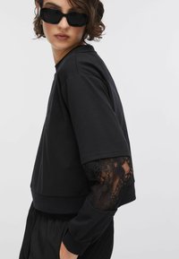 Pull-over noir avec encolure ronde, manches courtes et accents en dentelle sur les poignets. Tissu doux avec une coupe décontractée.