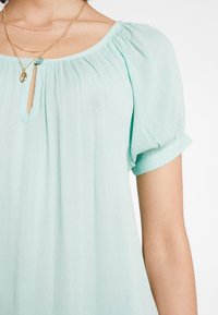 Blouse bleu-vert clair avec de courtes manches bouffantes, dotée d'un décolleté en keyhole et d'un tissu doux et texturé. Les manches roulées ajoutent une touche décontractée.
