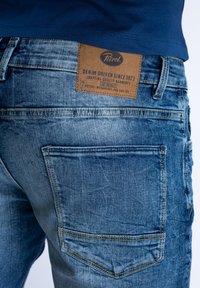 Wyczernione niebieskie dżinsy o zużytej teksturze, z brązową skórzaną metką z napisem "Petrol" oraz dwoma tylnymi kieszeniami.