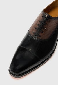 Chaussure de ville en cuir noir avec un bout pointu, présentant un dessus en cuir marron texturé et un laçage complexe. Fini lisse et design élégant.