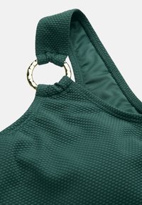 Mørkegrønt tekstureret stofrem med en rund guldfarvet metalringdetalje på et stykke tøj, vist mod en ensfarvet baggrund.