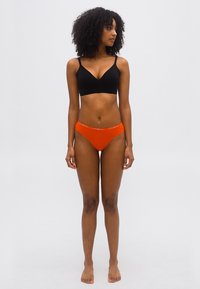 Νεαρή γυναίκα με κατσαρά μαλλιά, φορώντας μαύρο σουτιέν και πορτοκαλί εσώρουχα, όρθια ξυπόλητη μπροστά σε λευκό απλό φόντο, κοιτάζοντας προς τα πλάγια.