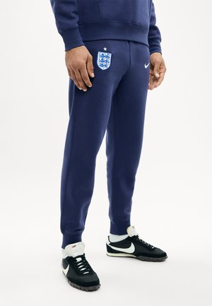 Personne portant un pantalon de survêtement bleu marine de l'équipe de football d'Angleterre et des baskets Nike noires avec des logos swoosh blancs, debout sur un fond blanc.