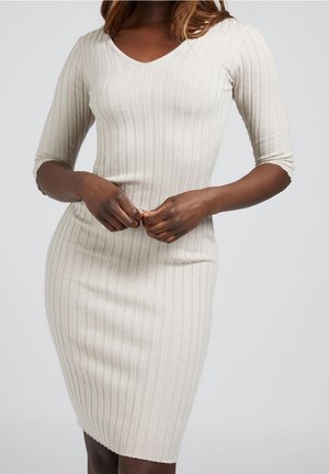 Kobieta ubrana w dopasowaną, beżową sukienkę z prążkowanego dzianiny z rękawami do łokcia, stojąca z rękami złożonymi na wysokości talii.