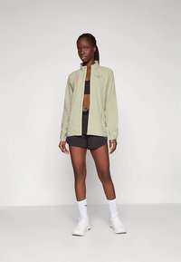Veste de sport vert clair avec une fermeture éclair à l'avant, des manches longues et un logo. Associée à un crop top noir et un short, portée avec des baskets blanches.
