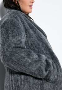Abrigo de piel sintética gris con una superficie suave y texturizada. Presenta una silueta relajada y mangas oversized, acentuadas por bolsillos laterales.