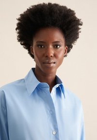 Camicia button-up azzurro chiaro con un ampio colletto, texture liscia e maniche lunghe. Presenta un singolo bottone vicino al colletto sul lato sinistro.