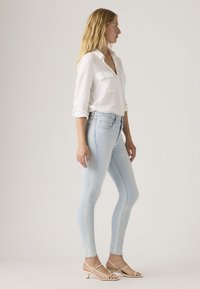 Γυναίκα που στέκεται πλάγια φορώντας λευκό πουκάμισο με μακριά μανίκια, ανοιχτό μπλε στενά τζιν και μπεζ σανδάλια με χαμηλό τακούνι και λουράκια σε απλό φόντο.