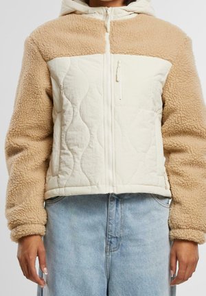 Beige sherpa fleece jas met crèmekleurig doorgestikt voorpaneel, ritssluiting, ritssluiting op de borstzak, gedragen met lichtblauwe spijkerbroek.