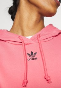 Zbliżenie na szyję osoby oraz różową bluzę Adidas z kapturem, z troczkami i czarnym logo na piersi, na białym tle.