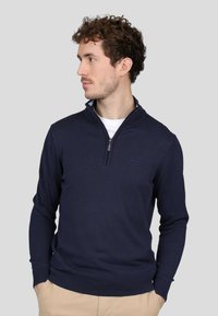 Pull à col zippé bleu marine en tissu tricoté. Il présente des manches longues, des poignets et un ourlet côtelés, ainsi qu'un discret logo sur la poitrine.