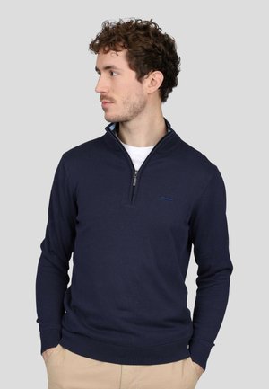 Pull à col zippé bleu marine en tissu tricoté. Il présente des manches longues, des poignets et un ourlet côtelés, ainsi qu'un discret logo sur la poitrine.