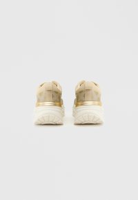 Scarpe da ginnastica beige con un accento oro metallico, tomaia in tessuto testurizzato e tallone imbottito; progettate per comfort e supporto.