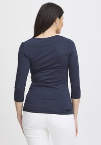 Blu închis, bluză cambrată cu mâneci trei sferturi, realizată dintr-un material moale. Prezintă un decolteu rotund și o textură netedă. Vedere din spate.