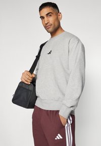 Sweatshirt cinza com punhos canelados, logotipo preto no peito. Calças bordô com riscas brancas. Saco preto com alça ajustável.