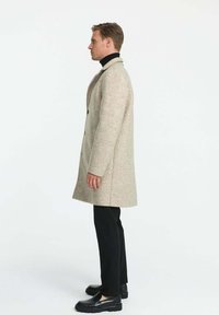 Bežový kabát z vlnenej zmesi s úzkym strihom, zapínanie na dva gombíky a bočné vrecká, skombinovaný s čiernymi nohavicami a topánkami.
