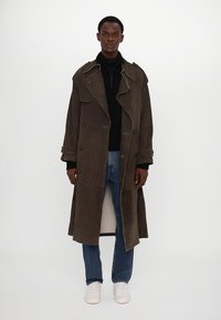 Langer brauner Wildledermantel mit großen Revers, doppelten Knöpfen, seitlichen Taschen und einer entspannten Passform. Getragen über einem schwarzen Rollkragenpullover und Jeans.