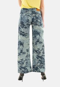 Jeans de pierna ancha en denim azul claro con un detallado estampado botánico. Presentan una textura suave y un parche trasero de cuero marrón.