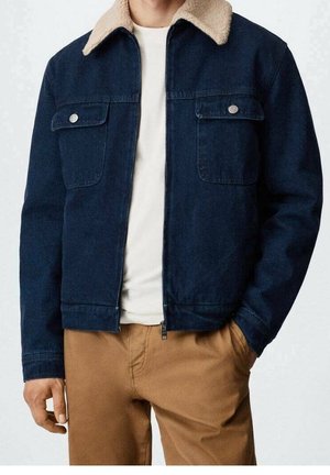 Man die een marineblauwe spijkerjas draagt met een beige fleecekraag, wit shirt en lichtbruine broek, staand met één hand in de zak.