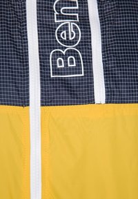 Giacca colorata con una parte superiore blu scuro a motivo a griglia, una parte inferiore gialla e una zip bianca con elementi di branding.