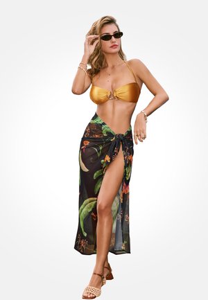 Kobieta w złotym topie od bikini, czarnym kwiecistym sarongu związanym w talii, okularach przeciwsłonecznych, bransoletkach i beżowych sandałach na obcasie, pozująca z ręką na okularach przeciwsłonecznych.