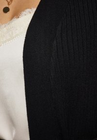 Musta ribattu neulecardigan tiiviillä leikkauksella, jossa on tekstuuri pinta ja valkoinen pitsireunainen paita vilkkuu sen alta.