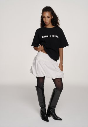 Frau mit lockigem Haar, die ein schwarzes T-Shirt mit der Aufschrift "GIRL'S GIRL", einen weißen Puffrock, schwarze Strumpfhosen und kniehohe schwarze Stiefel trägt und vor einem einfarbigen Hintergrund posiert.