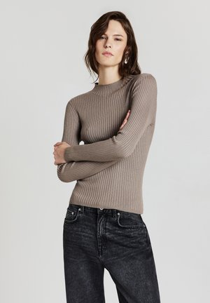 Beige Rippenstrickpullover mit einem Stehkragen und langen Ärmeln, kombiniert mit dunklen, verwaschenen Denim-Jeans. Schlichtes und figurbetontes Design.