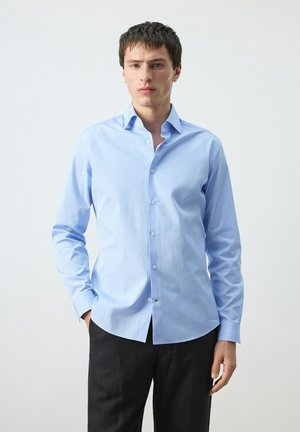 Junger Mann trägt ein hellblaues, geknöpftes Hemd und schwarze Hose, steht mit einer Hand in der Tasche vor einem einfarbigen Hintergrund.
