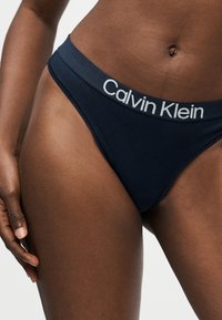 Κοντινό πλάνο ενός ατόμου που φοράει μαύρα εσώρουχα Calvin Klein, εμφανίζοντας το λογότυπο στη ζώνη μέσης και το μέσο του σώματος με το χέρι να ακουμπάει στο μηρό.