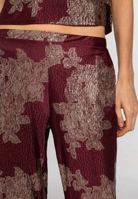 Acercamiento de una persona que lleva pantalones burdeos y una blusa con patrones florales plateados texturizados, con la mano descansando cerca del muslo.