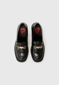 Mocassins en cuir noir à bout rond, ornés d'une chaîne en or. Semelle intérieure en forme de cœur rouge avec l'inscription "LOVE MOSCHINO". Texture lisse.