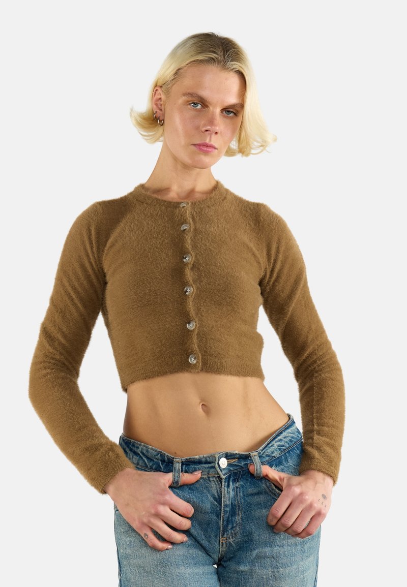 Junge Frau mit kurzen blonden Haaren, die einen braunen, flauschigen, geknöpften Cropped-Top und eine blaue Jeans trägt, posiert mit den Händen in der Nähe der Taschen.