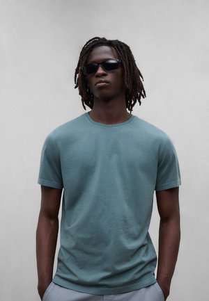 Ung mann med skulderlange dreadlocks iført svarte solbriller og en turkis t-skjorte med korte ermer, stående mot en ensfarget grå bakgrunn.