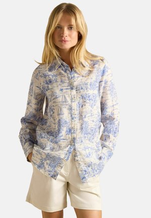 Blonde woman wearing a buttoned, long-sleeve shirt with blue scenic prints and beige shorts, standing with hands in pockets.