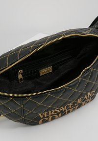 Black quilted bag with gold hardware and text "VERSACE JEANS COUTURE." Interior includes pocket with zipper, lined in black fabric.