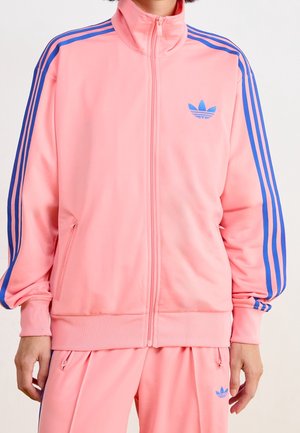 Personne portant une veste et un pantalon de survêtement Adidas rose avec des rayures et des logos bleus, debout les mains détendues le long du corps.