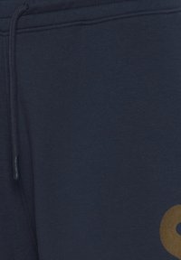 Marineblå bomulls joggebukser med en snor i midjen og en stor gul logodetalj. Myk tekstur med en jevn overflate.