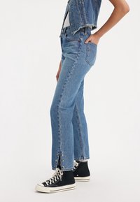 Pantalones vaqueros azules de mezclilla con un corte recto, con dobladillo sin rematar y detalles de estrellas en las aberturas laterales, combinados con zapatillas negras de caña alta.