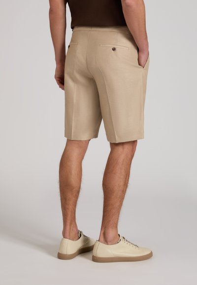 Man wearing beige tailored shorts and cream sneakers, standing with hands in pockets, shown from the back against a plain background.
