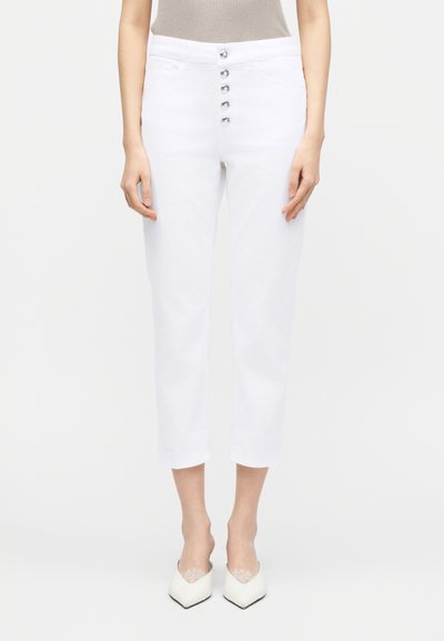 Γυναίκα που φοράει λευκό ψηλόμεσο κοντό παντελόνι με πέντε ασημένια κουμπιά στο μπροστινό μέρος και λευκά παπούτσια με μυτερή μύτη σε λευκό φόντο.