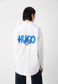 Hvit langermet skjorte med blå spraymalt "HUGO"-logo på baksiden, med buet hem og knappmansjetter. Glatt stofftekstur.