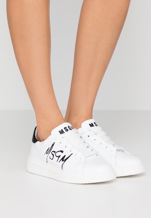 Witte MSGM lage sneakers met een zwart logo op de zijkant en tong, gedragen op blote voeten tegen een effen lichte achtergrond.