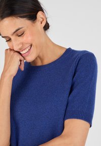 Pull à manches courtes en tricot de couleur bleu vif. Il présente un col rond et une texture délicate avec des variations subtiles de teinte.