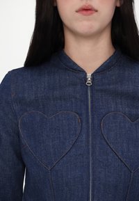 Jeans jakna z zadrgo spredaj, okroglo ovratnico in žepoma v obliki srca. Temno modra tkanina z oranžnimi šivi.