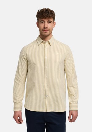 Jonge man die een beige en witte, verticaal gestreepte, overhemd met lange mouwen en donkere broek draagt, staand tegen een effen achtergrond.