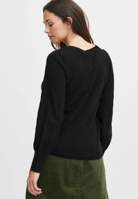 Schwarzer Strukturpullover mit einem Rautenmuster, gerippten Bündchen und einem V-Ausschnitt auf der Rückseite, kombiniert mit einem grünen Cordrock.