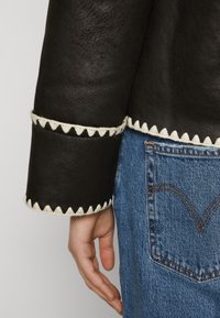 Main reposée à côté de la poche d'un jean en denim bleu, portant une veste en cuir brun foncé avec une bordure triangulaire blanche sur les manches et l'ourlet.