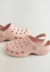 Ružové dreváky vyrobené z ľahkého plastu, s viacerými kruhovými otvormi na ventiláciu, s designom na obúvanie a so zadným popruhom pre bezpečné uchytenie.