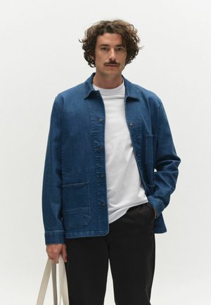 Mann mit lockigem Haar und Schnurrbart, der eine blaue Jeansjacke, ein weißes T-Shirt und schwarze Hosen trägt und eine beige Tragetasche vor weißem Hintergrund hält.
